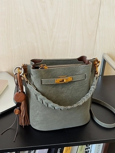 Minimalist grey-green bucket bag with golden twist lock and tassel charm on desk