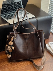 Large dark brown tote bag with woven strap and rabbit charm on a leather bench