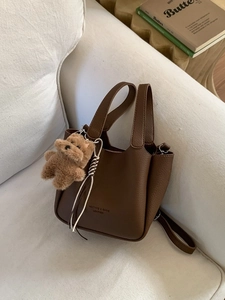 Deep brown pebble leather bucket bag with top handles and plush bear charm on white sofa
