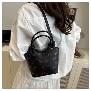 Detail of a black bucket bag with red cherry prints and slim shoulder strap on a model