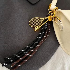 Detail van een gouden tennisracket hanger met geweven kwastjes aan een witte leren tas