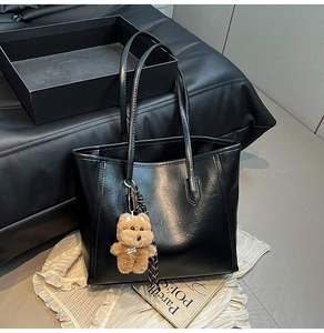 Detail of black faux leather tote bag with a brown plush bear charm on white cloth