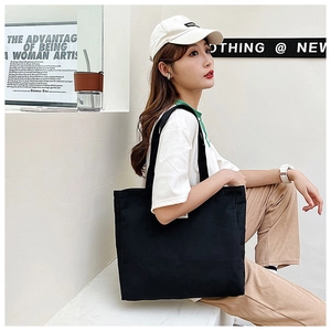 Detail of a black canvas tote bag with a white side label worn by a model in a minimal setting
