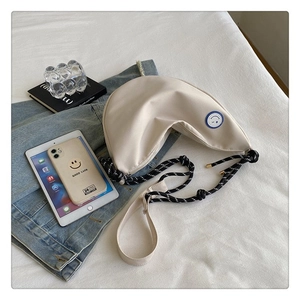 Détail du sac croissant en toile beige avec bandoulière tressée et patch smiley bleu