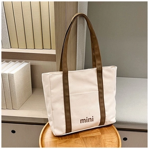 Vista detallada del bolso tote de lona crema con asas marrones y mini logo sobre un taburete