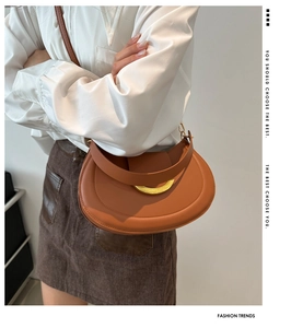 Detail of a brown leather saddle bag featuring a gold crescent buckle and slim shoulder strap