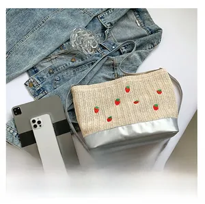 Detail of woven straw bucket bag featuring red strawberry embroidery and silver PU strap