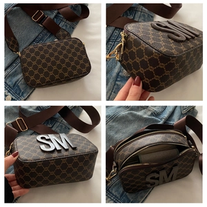 Detail of a brown monogram camera bag with SM logo, double zippers, and wide canvas strap