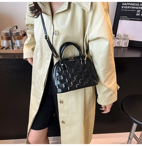 Fashionable black patent leather shell bag with woven texture worn by a model in a room