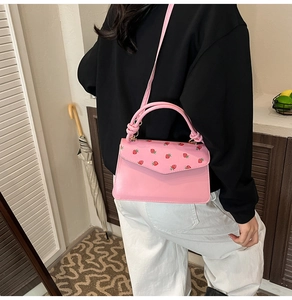 Pink crossbody bag featuring red strawberry prints and a woven handle in a cozy home setting