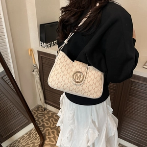 Beige Umhängetasche mit M-Muster und goldener Metallschnalle in wohnlicher Umgebung