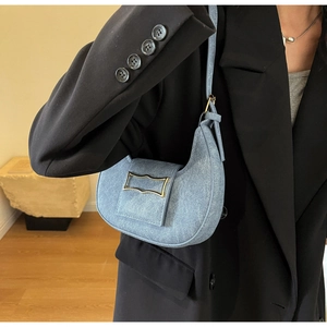 Détail d'un sac baguette en denim bleu avec boucle dorée porté sur un blazer noir chic