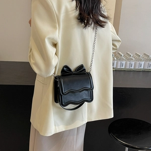 Chic black chain shoulder bag with large bow and wave flap, worn by a model in a modern room