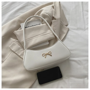 Detail of white half-moon bag with gold bow ornament next to a phone for size comparison