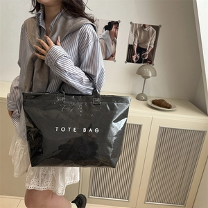 Glossy black tote bag with white TOTE BAG print held by model in minimalist room
