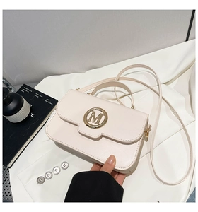 Détail d'un sac bandoulière en cuir beige avec un logo M métallique doré sur le rabat avant