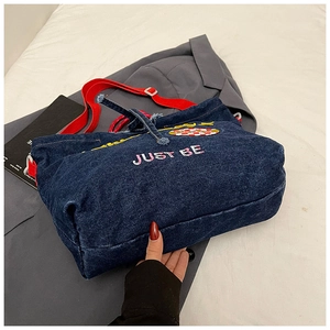 Detail of denim tote bag showing red shoulder strap and embroidered JUST BE lettering