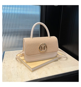 Détail du sac beige avec rabat tissé et boucle dorée en M posé sur un livre jaune