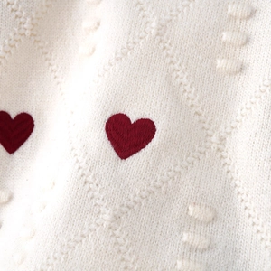 Nahaufnahme von cremefarbenem Strickpullover mit rotem Herz-Stickmuster und Rautenstruktur