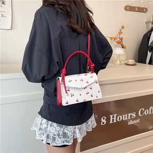 Elegant white crossbody bag with cherry prints and red handles shown in a cafe setting