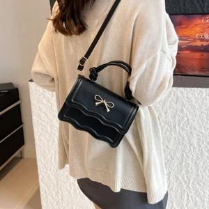 Elegant black handbag with wavy flap and gold bow detail worn as a crossbody bag