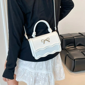 White mini handbag with wavy flap and gold bow accessory worn by model over black dress