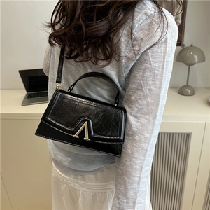 Chic black glossy leather shoulder bag with gold V clasp on model in minimalist room