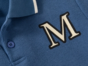 Macro detail of navy knit polo shirt with white striped collar and ivory letter M patch