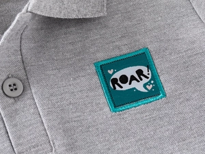 Detailed macro view of the grey pique cotton fabric and ROAR dinosaur patch on the polo shirt
