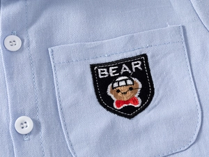 Detail of light blue cotton shirt featuring a white button and cartoon bear badge embroidery