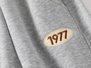 Détail macro du tissu gris chiné avec écusson brodé marron 1977 sur le pantalon