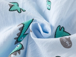 Close-up of soft light blue cotton fabric with green cartoon dinosaur print and Hello text