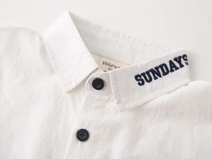 Detailed close-up of white cotton linen boy shirt featuring blue SUNDAYS collar embroidery