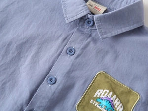 Detail of toddler blue cotton shirt featuring a dinosaur patch and size 90 label