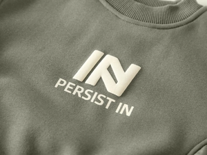 Detail of sage green sweatshirt with embossed white PERSIST IN logo and ribbed neckline