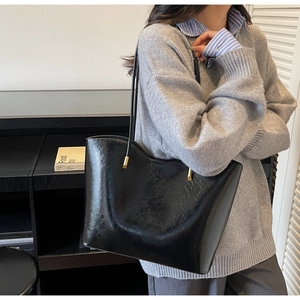 Detail of glossy black tote bag with gold metal hardware on model in grey sweater