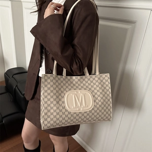 Elegant beige tote bag with M-pattern and shoulder strap shown in a modern lifestyle setting