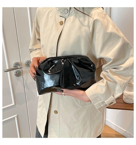 Detail of a black glossy pleated cloud clutch bag held by a model in a beige trench coat