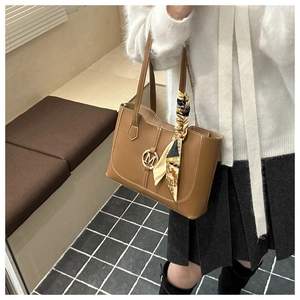 Close-up of caramel tote bag featuring a gold M-logo and a silk scarf tied to the handle