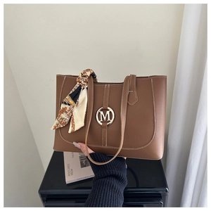 Detail of a brown leather tote bag with white stitching, metal M logo, and silk scarf wrap
