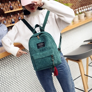 Dark green corduroy mini backpack with red tassel charm shown on model in modern cafe
