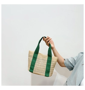 Detail of woven straw tote bag featuring contrast dark green fabric handles on white background