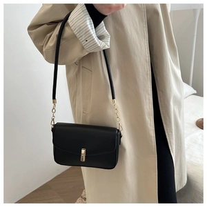 Detail of a matte black square crossbody bag with gold buckle on a beige trench coat