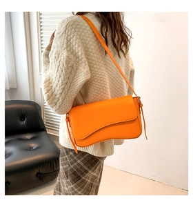 Vibrant orange shoulder bag with asymmetric flap shown on a model in a cozy setting