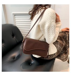 Detail of dark brown shoulder bag with wavy flap shown on a model sitting on a chair