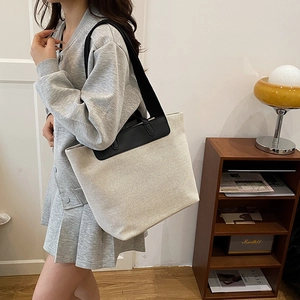 Large beige linen tote bag with black leather straps worn by model in a cozy room