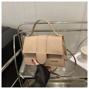Détail du sac beige à rabat avec surpiqûres verticales et logo doré tenu à la main