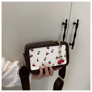 Detail of white crossbody bag with red cherry print, brown trim and wide shoulder strap
