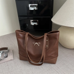 Minimalist large brown leather tote bag with elegant golden bow charm at 45-degree angle