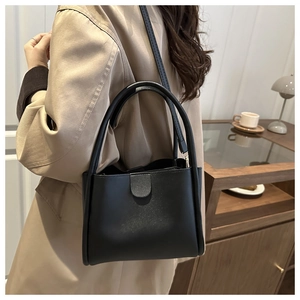 Minimalist black bucket bag shown as a crossbody on a trench coat in a warm cafe setting